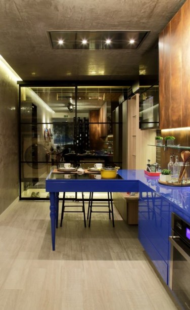 CASACOR Alagoas 2015. Cozinha com Área de Serviço e Varanda Gourmet - Amanda Vilela e Carol Couto. O ambiente de 43m² é paginado com porcelanato que lembra madeira e recebe armários suspensos com diferentes alturas. Na parede e no teto foi aplicada a textura stucco veneziano, de efeito marmorizado. Com essa base, os elementos de destaque passaram a ser a bancada e os armários laqueados no tom azul real. Portas de correr em vidro Reflecta separam o ambiente da área de serviço e recebem uma TV embutida.