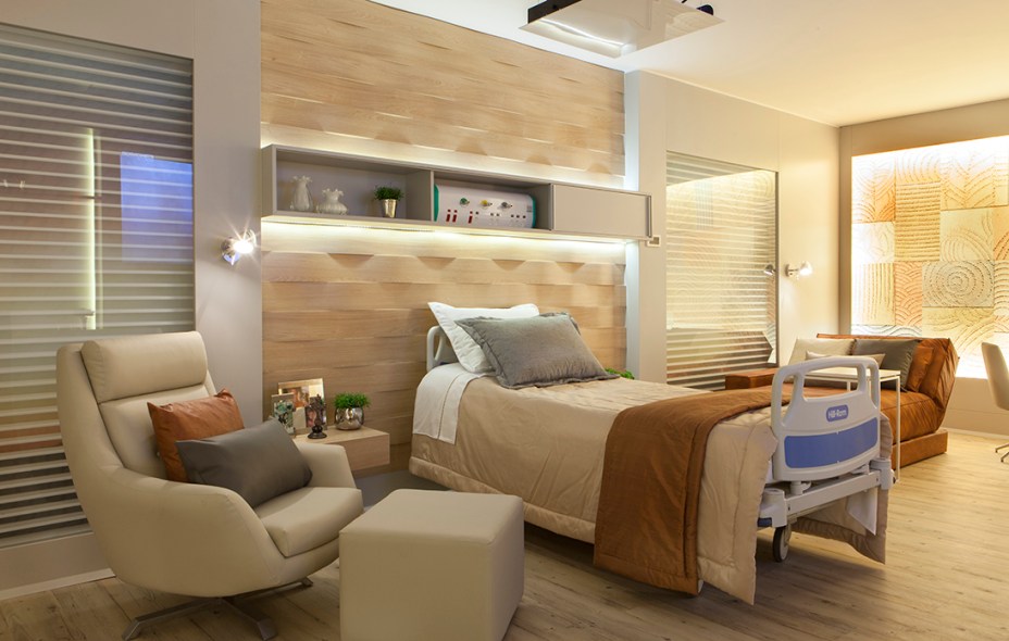 Quarto de Hospital - Alessandra Guidoni. A atmosfera é residencial e com detalhes que remetem à hotelaria de luxo, para humanizar o espaço e deixar o paciente à vontade. A iluminação é indireta e os revestimentos adicionam beleza e funcionalidade. Há até espaço para a arte, evidente no grande painel em acrílico.