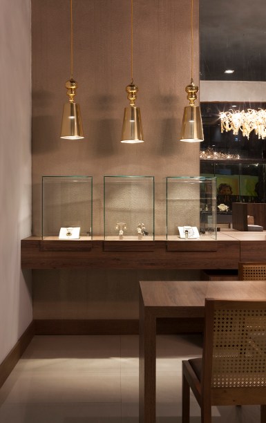 Joalheria - Luciana Schaeffer e Wilber Soares. A exposição das jóias é pensada para torná-las objetos de desejo, sejam protegidas em redomas de vidro ou em uma vitrine feita em pedra. Uma luminária feita artesanalmente é destaque, assim como os pendentes dourados que enobrecem ainda mais a composição.