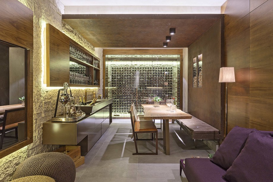 Sala de Vinho - Sílvia Carvalho. Armazenar, degustar e compartilhar são verbos conjugados no espaço que valoriza a experiência com o vinho. A adega climatizada é o foco e, perto dali, a mesa acomoda oito pessoas. Uma bancada centraliza as funções de pia, champanheira e cooktop, além de servir de apoio para limpeza de taças e montagem dos pratos. A área é demarcada pela parede de pedras irregulares, com textura acentuada pela luz do painel em madeira.