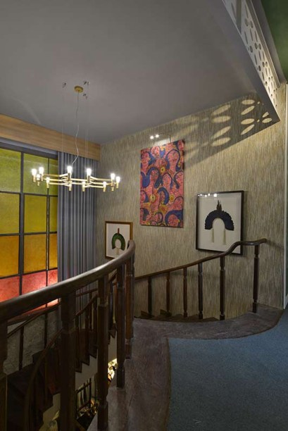 Hall da Escada da Família - Roger Monte. Os vitrais nas cores do pôr-do-sol são destaque no ambiente, que convoca uma série de memórias afetivas ligadas à arquitetura brasileira na escada vintage, nos objetos garimpados e nas artes plásticas com curadoria de Aldonso Palácio. A cortina foi produzida em linho, com técnica de alfaiataria. O piso recebeu uma camada de quartzito Perla Santana, extraído no próprio estado, enquanto os revestimentos das paredes combinam pedras, um madeirado de Peroba e papel de parede em palha natural.