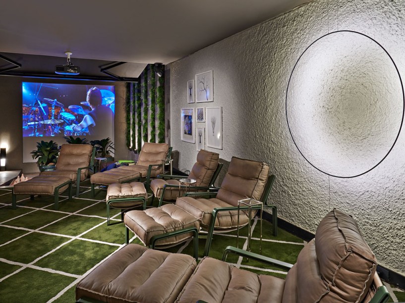 Home Theater - Adriana Mundim e Fernando Rocha Galvão dividiram os 58m² em ambientes externo e interno. Neste, diversas poltronas individuais em couro fendi e estrutura verde são protagonistas, sobre o tapete geométrico desenhado pelos profissionais - e que contribui para a acústica. O teto é uma laje lisa e livre de forro, que recebeu uma pintura na tonalidade cinza. Na iluminação, o destaque fica para as peças Catellani Smith que tratam a luz como escultura, com uso do LED. É possível ver o telão, que fica na parte descoberta, próximo ao painel com pedras Hitam Palimanam intercaladas com o jardim vertical.