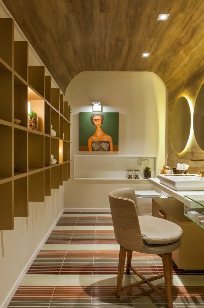 CASACOR Pernambuco 2016. Lavabo da Jovem - Cynthia Costa. O painel em madeira envolve teto e paredes para marcar um cenário aconchegante. Nele, o espaço da maquiagem é garantido, na mesma bancada da cuba com metais dourados. Os modulados seguem a sofisticação, com o acabamento high gloss perolizado na cor ouro. Para arrematar, tapete exclusivo em pastilhas de vidro.