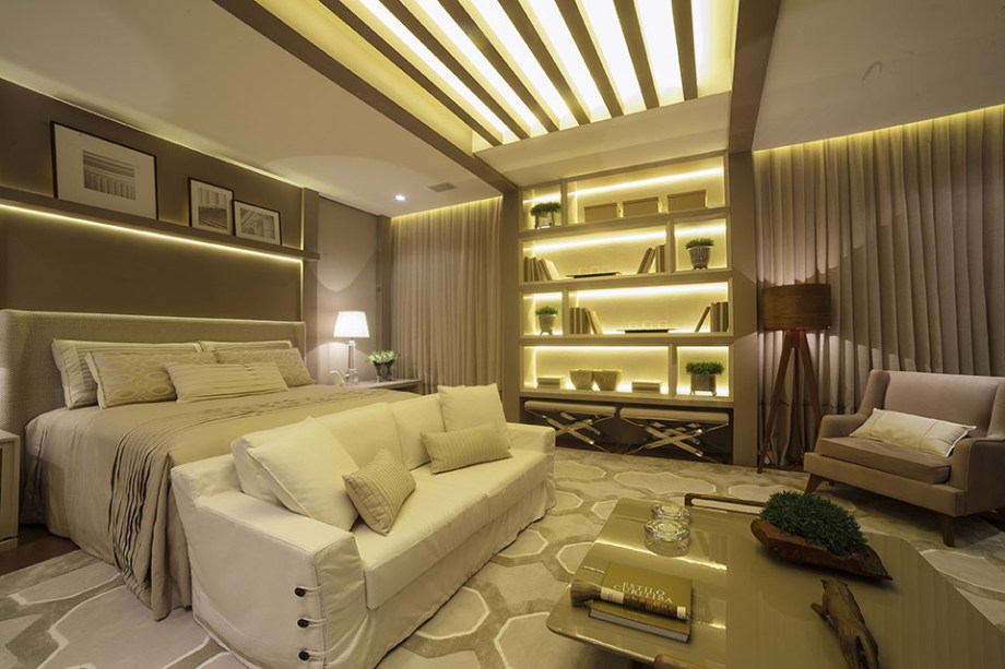 Quarto do Casal - Viviane Loyola. O quarto inclui um ambiente de estar, e o projeto luminotécnico atende a essas duas cenas, priorizando efeitos de luz indiretos. Ele valoriza alguns detalhes, como as réguas de madeira no teto, que prolongam as linhas da estante embutida. Tons de creme e fendi se revezam, para criar uma atmosfera de tranquilidade nestes 40 m².
