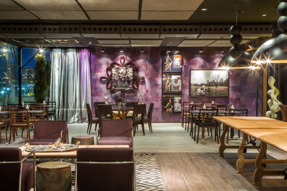 Restaurante Casa Cor - O ambiente de Cristina Côrtes é pautado pelos tons de cimento queimado, que interagem com a aparência de madeiras de demolição no piso vinílico. Vigas metálicas com pintura em aço corten e dutos aparentes desenham o espaço. Paredes laterais ganharam revestimento em papel (Guilha) com padronagem patchwork na cor berinjela. As mesas combinam aço, vidro e couro, acompanhadas de cadeiras e bancos estofados em linho e seda. Duas mesas de demolição, assinadas pela dupla Laura e Roberta Pederneiras, se destinam ao bufê. Em primeiro plano, você vê também o lounge de espera, com poltronas em aço revestidas em seda e mesa de centro de demolição. Os pendentes em forma de móbile são da designer Luciana Martins.
