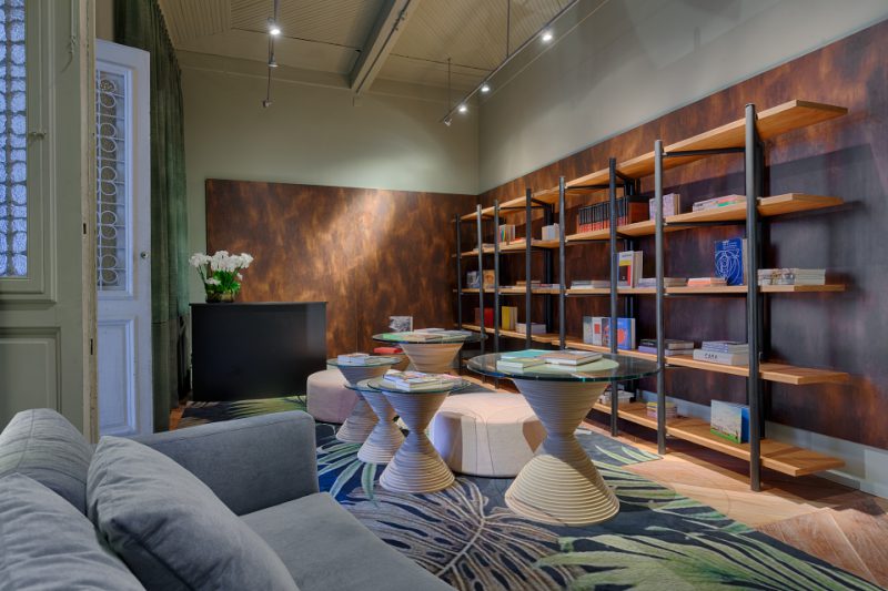 Livraria e Varanda - Camila Ferreira. <span><strong>Mesas centrais</strong>: A estrutura redonda foi criada com a intenção de retirar a rigidez do espaço e oferecer mais movimento e leveza. Os pés foram concebidos em MDF cru de vários diâmetros diferentes, o que causa a sensação de escalonamento. A cor neutra escolhida para os pés foi pensada para não brigar com as madeiras originais existentes no ambiente e combinar com o tapete. </span><span><strong>Estantes:</strong> Pelo fato da mostra habitar uma construção tombada e não suportar fixação na parede, a estante foi uma ótima solução. Foram criados cavaletes de metalon pintados e as prateleiras em lâmina de freijó natural bastante robustas, para sustentar o peso dos livros.</span>