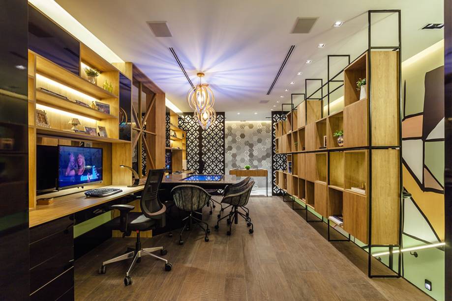 CASACOR Rio Grande do Sul: Home Office do Casal - Carol Fernandes Heck e Gustavo Heck. Para o espaço, os profissionais pensaram em duas estações de trabalho foram organizadas lado a lado e, entre elas, foi posicionada a mesa de reuniões com TV touch. A marcenaria utiliza melamina Carvalho. A estante ortogonal de ferro preenchida com módulos quadrados acrescenta volume, atua como guarda-corpo da escada e permite efeitos de iluminação.