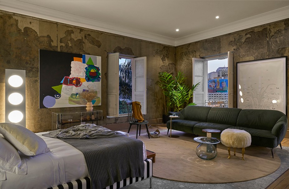 CASACOR Minas Gerais. Loft - Ana Bahia. O que é original do casarão está impresso no pé direito alto e nas paredes descascadas, valorizadas com acabamento em verniz. Para trazer mais conforto, o toque delicado do sofá em veludo e da cama revestida em tecido listrado bicolor. Peças soltas no mobiliário conferem um toque despojado e delicado, explorando formas e transparências.