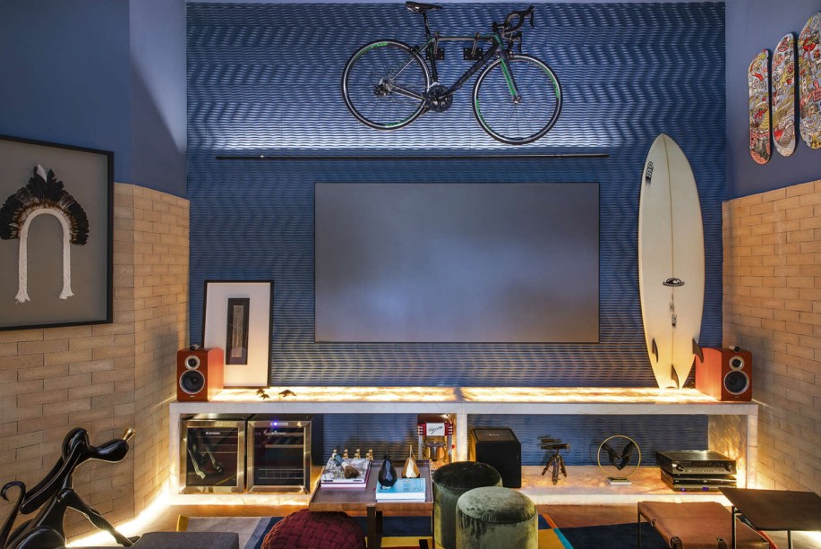 Patrícia Makhoul, Lucas Martinelli e Áurea Degani - Sala da Família. Para uma família com história para contar, um espaço descontraído. Um mood que permite transformar a bicicleta em peça de decoração, disposta junto com objetos trazidos de viagens memoráveis. O telão reúne todo mundo para assistir a uma série, e a luz indireta acolhe, partindo das laterais e do móvel em pedra. Um detalhe: o piso original da casa, de pau brasil, certamente foi mantido.