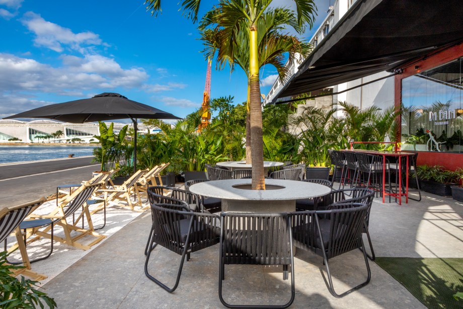 Isabella Lucena - Restaurante da Baía. O estilo art-déco, com suas formas circulares, foi a grande inspiração para a criação do restaurante desta edição. Ele aparece nos elementos vazados em formato de arco que fazem as vezes de divisória, nas luminárias pendentes e em algumas peças de mobiliário. O pilar no centro do espaço foi transformado numa espécie de ilha verde com duas enormes palmeiras de quatro metros de altura e muitas plantas em todo o seu entorno. O lounge também ganhou um formato diferente: uma arquibancada em L oferece um jeito mais descontraído de esperar uma mesa e apreciar a vista para a Baía.