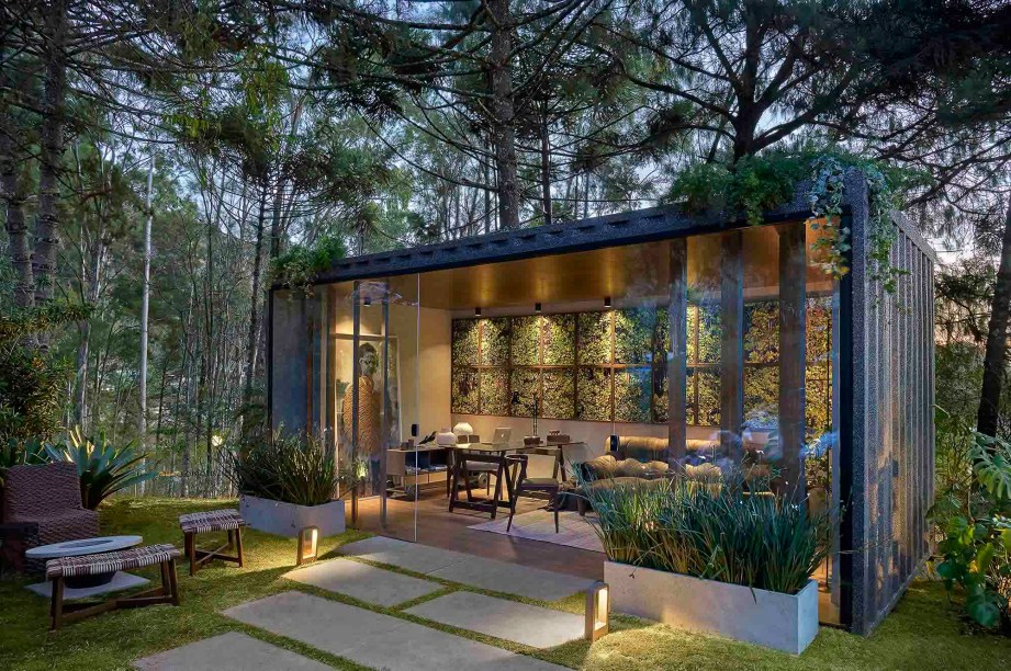 CASACOR Minas Gerais 2019. Escritório do Jardim - Marina Diniz e Paula Guimarães. Um home office em pleno quintal. O contêiner foi revestido na parte externa com um ecogranito resistente e de fácil aplicação. A pedra sabão faz o caminho pelo gramado até o ambiente pontuado por poltronas e pufes em corda náutica e mesas de apoio em fórmica, para reuniões informais ou apenas relaxar. Internamente, o design do mobiliário sobressai, com peças de Sérgio Rodrigues, Jorge Zalzupin e Jader Almeida. As 22 telas de Alessandra Rehder formam uma espécie de janela.