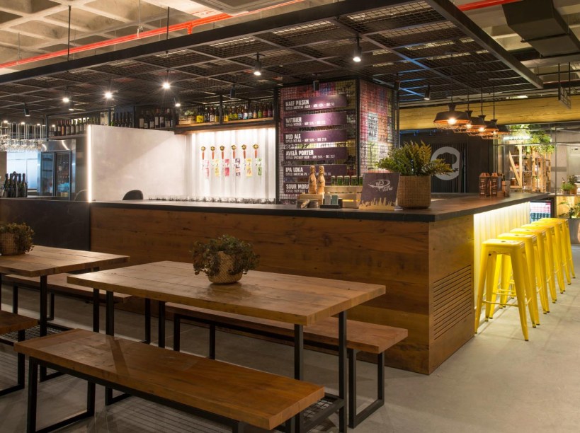 CASACOR Santa Catarina | Florianópolis 2019. Beer Bar - Fabio Bubniak e Ana Carolina Melo. O estilo industrial conceitua o projeto e balizou a escolha de acabamentos que exibem os materiais em estado bruto. Entre eles, a bancada em pedra natural e a chapa metálica de contêiner na frente do balcão. A madeira de demolição aparece em painéis e estantes, junto com quadros metálicos e telas aramadas, adicionando possibilidades para expor produtos.