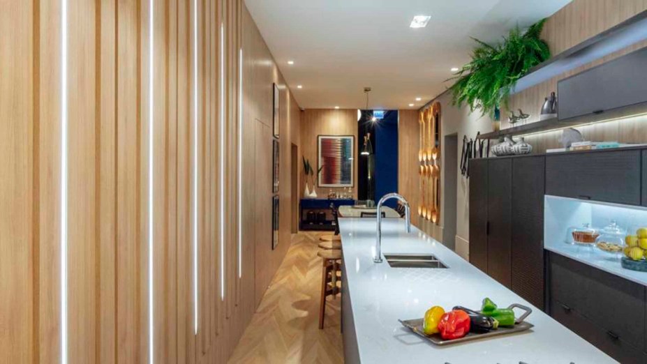 CASACOR Rio Grande do Sul 2019. Linear House - Renato Ferrari. A linearidade foi a solução encontrada para organizar todos os espaços em uma casa de 3 metros de largura por 18 de comprimento, totalizando 56 m². Para quebrar o padrão, a cozinha ficou entre a sala de jantar e o living, afirmando-se como o coração da casa. Para otimizar ainda mais a área, a iluminação foi distribuída em rasgos nas paredes, como um elemento geométrico adicional. O piso foi elaborado em Tauari de reflorestamento.