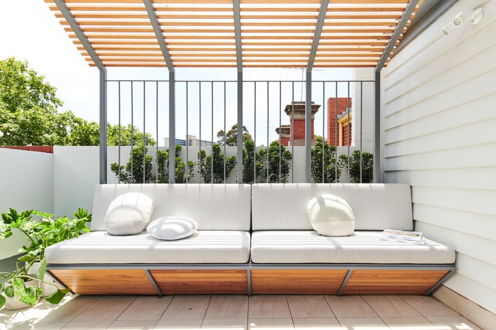 casa-australiana-com-mobilia-embutida-casacor-9 Banco ao ar livre, com suporte de madeira e estofado em branco, coberto por um gazebo de madeira. Ao fundo, plantar verdes.