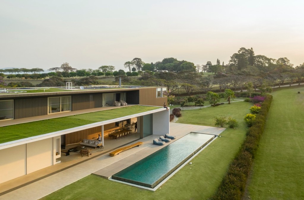 Visão superior do Telhado Verde, na diagonal, com uma pessoa sentada na beirada. Abaixo, no primeiro piso, uma piscina, com gramado em volta e no interior, uma grande sala de estar.