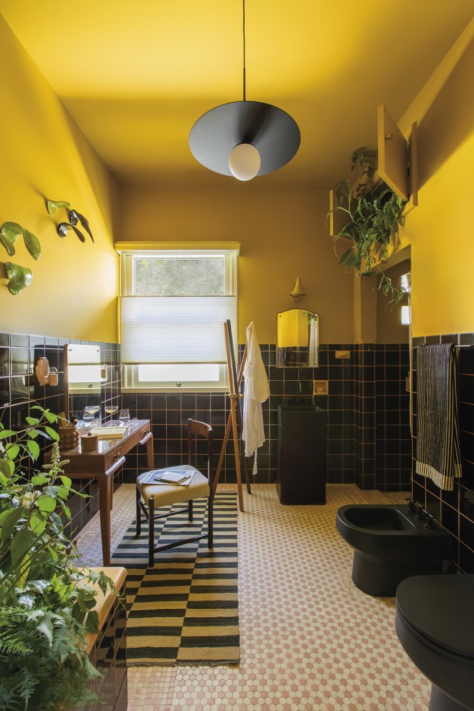 Isabela Bethonico e Studio Tertúlia - Quarto de banho: Rosa ou Qualquer coisa que faça quase sentir. Projeto da CASACOR Minas Gerais 2023. Na foto, banheiro com parede amarela, louças pretas, penteadeira e plantas.