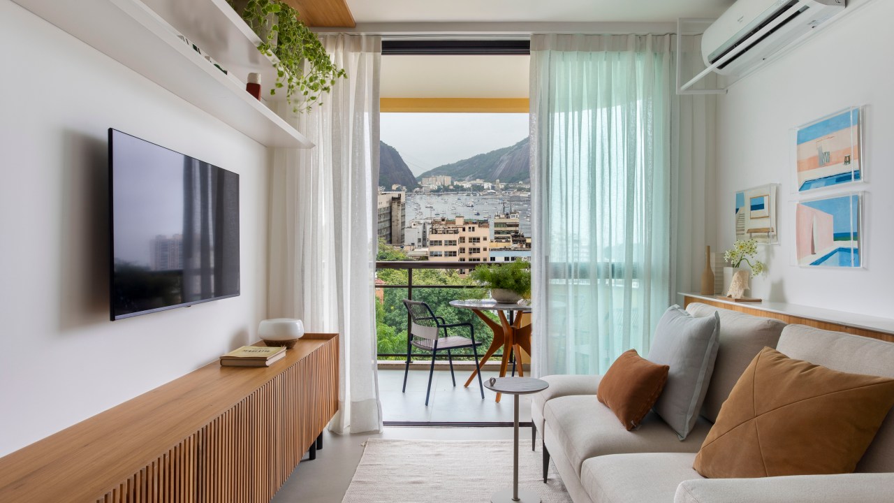 Branco e madeira criam décor cozy em apartamento de 70 m². Projeto de Rafael Ramos. Na foto, sala com tv, aparador ripado e quadros.