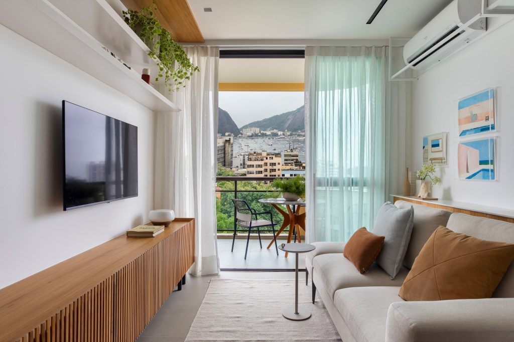 Branco e madeira criam décor cozy em apartamento de 70 m². Projeto de Rafael Ramos. Na foto, sala com tv, aparador ripado e quadros.