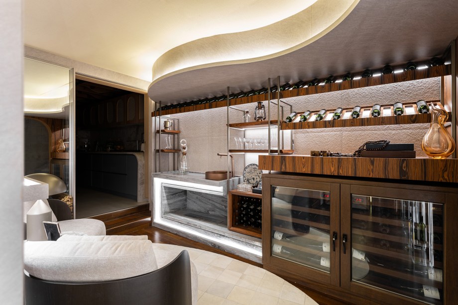 <b>Janaina Marques e Camila Kubota - Adega Residencial. </b>Além de ser uma adega funcional, este espaço proporciona uma experiência imersiva para os apreciadores da enologia. Com uma área climatizada que segue todas as exigências técnicas para a conservação dos vinhos, o ambiente inclui uma parede dedicada à exposição de rótulos especiais, uma bancada equipada com cuba e apoio para o manuseio das garrafas, e poltronas para degustação das bebidas. As arquitetas utilizam a marcenaria para criar um fluxo que guia o visitante até o ápice do projeto, onde desenham uma tela tensionada com padrões que remetem aos formatos das garrafas. Na lateral, o painel com prateleiras de vidro destaca ainda mais os rótulos especiais.